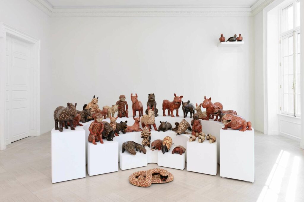 About 30 ceramic sculptures of various animals are positioned on staggered stages, shaped to evoke an arena. On the floor sits a ceramic sculpture of a coiled snake. In the upper right corner of the image a host of birds are perched on a shelf attached to the wall. 