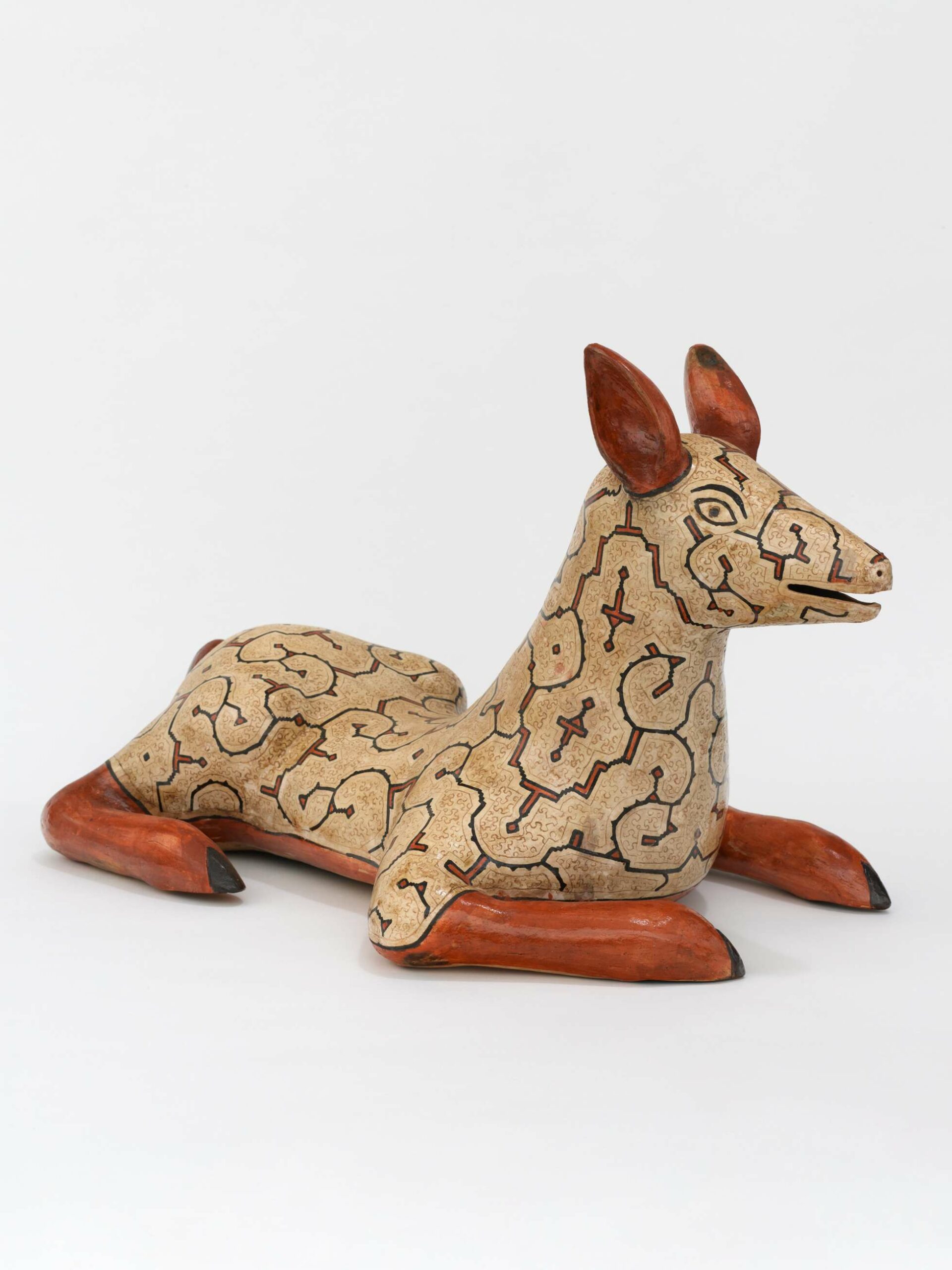 A ceramic sculpture of a deer lying on its belly, its four legs splayed. The deer has burnt red legs and ears, and it's body, rendered in beige, is covered in brown geometric patterns.
