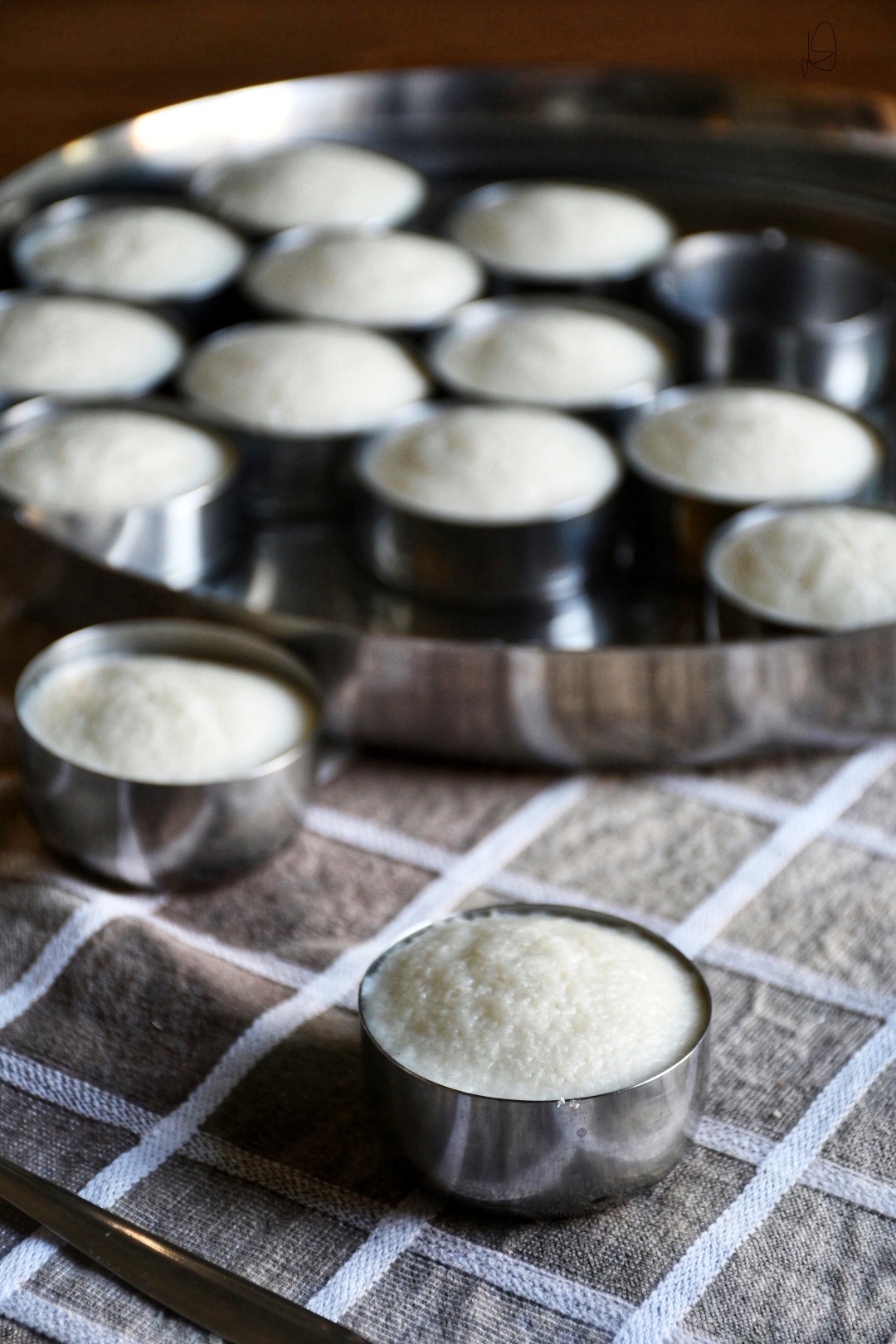 A tray of fluffy white idli are in individual steel containers on a beige and white checked tabelcloth.