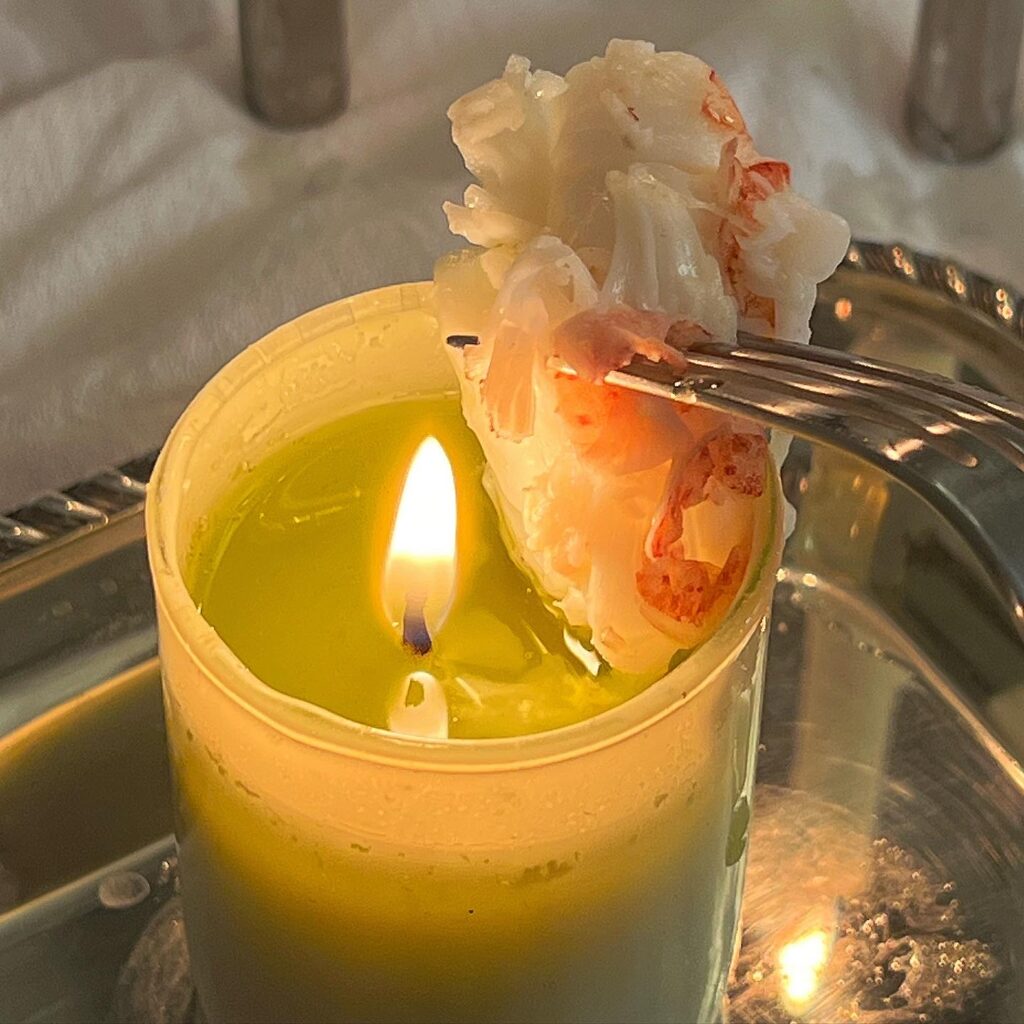 A lit candle, with a pool of wax at the top, a chunk of lobster tail is held aloft, dipped in the butter.