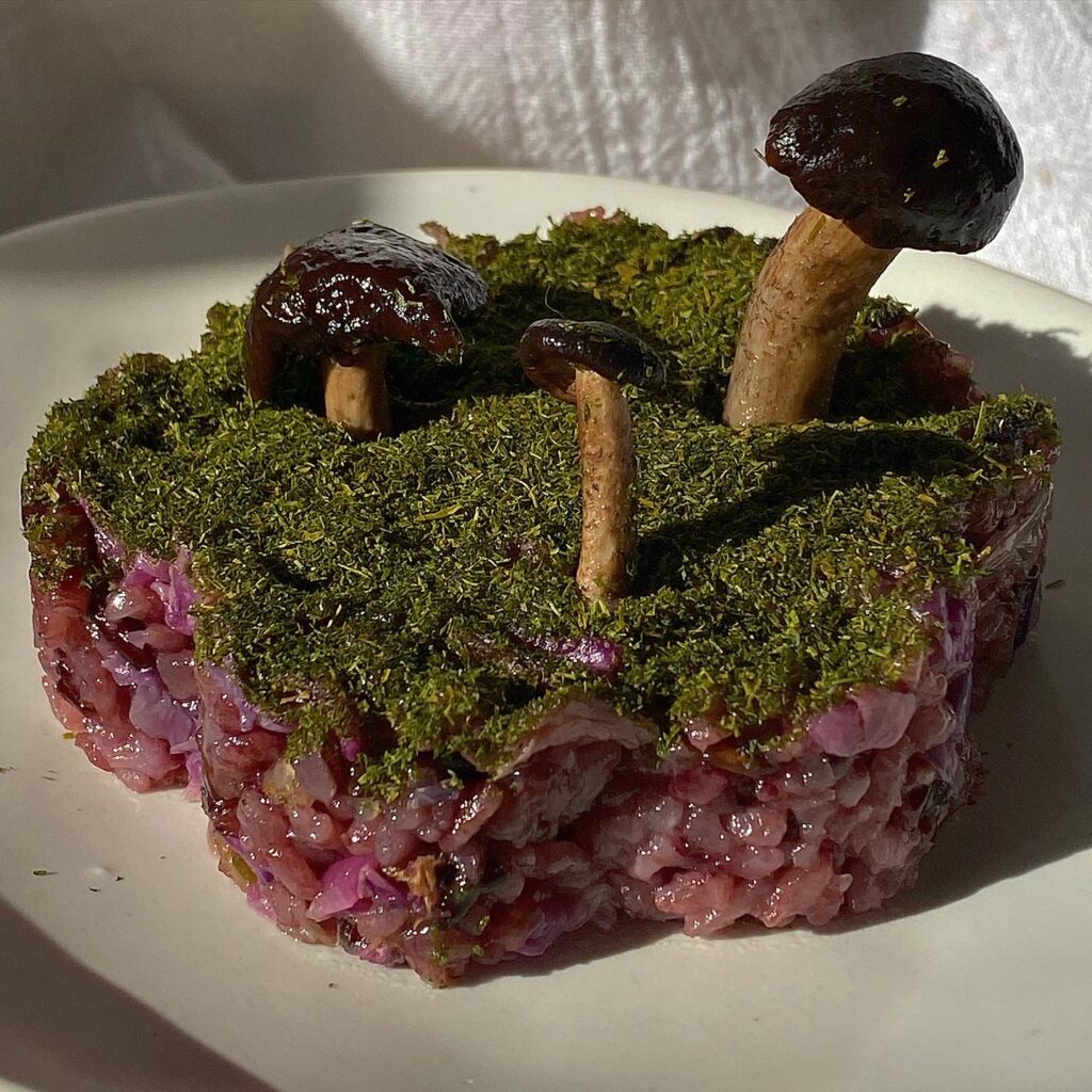 Purple rice fashioned in the shape of a flower, the top of the rice flower is covered in a layer of flaky seaweed, three mushrooms of varying sizes are positioned on top, emulating a forest.