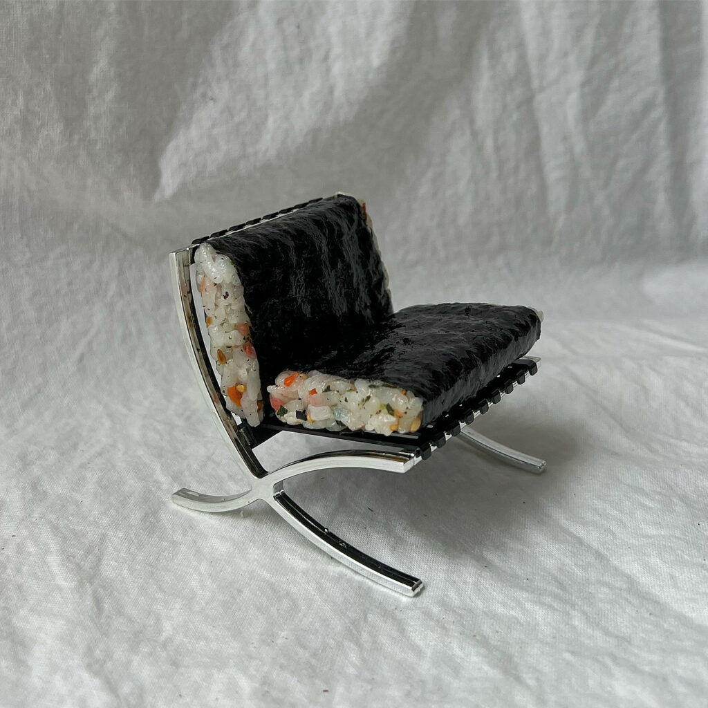A miniature metal chair frame against a white sheet background. Instead of chair cushions, there are onigiri fashioned in the shape of cushions. 