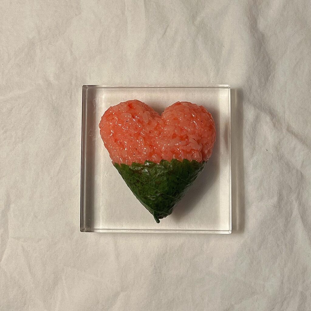 A heart-shaped rice ball lies on a glass plate on a white tablecloth background. The bottom half of the heart rice ball is wrapped in seaweed.