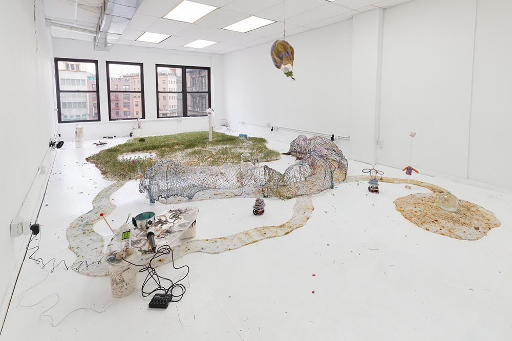 A room with white floors and walls is covered with a patch of grass interspersed with small circuits, miniature trellises made of yarn and wire, and small pools of plastic. A cloth hot air balloon floats above the miniature landscape