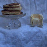A miniaturized butter model of an armchair sits on a white backgroud next to a clear glass platter with a stack of pancakes on top