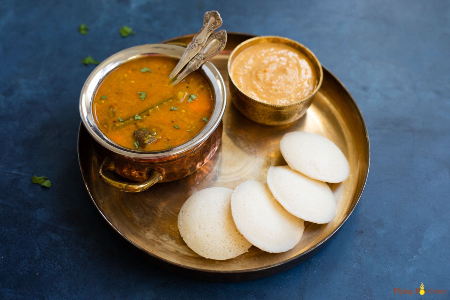 A plate of four idli is served with a bowl of sambhar and chutney.
