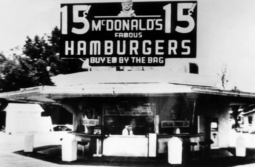 A black and white photo of a fastfood restaurant, a billboard reading McDonald's Famous Hamburgers 15 cents hangs above the building.