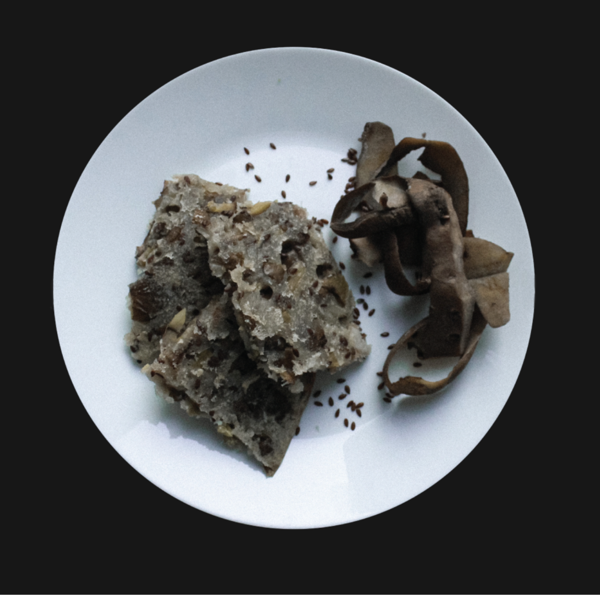A brown-gray flatbread broken into three pieces on a plate. Garnished with dried grass and scattered flax seed.