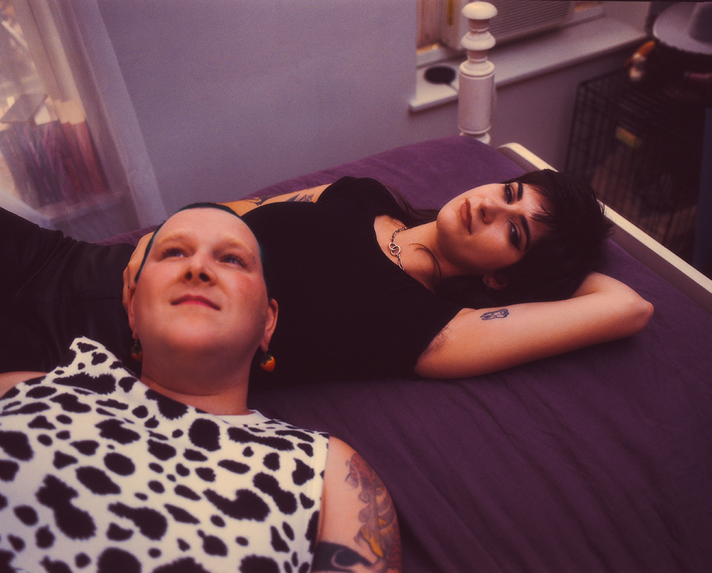 One person wearing a cow print tank top lies across the stomach of another individual reclining in a black t-shirt with their arm behind their head. They are both on a four post bed with eggplant purple sheets.