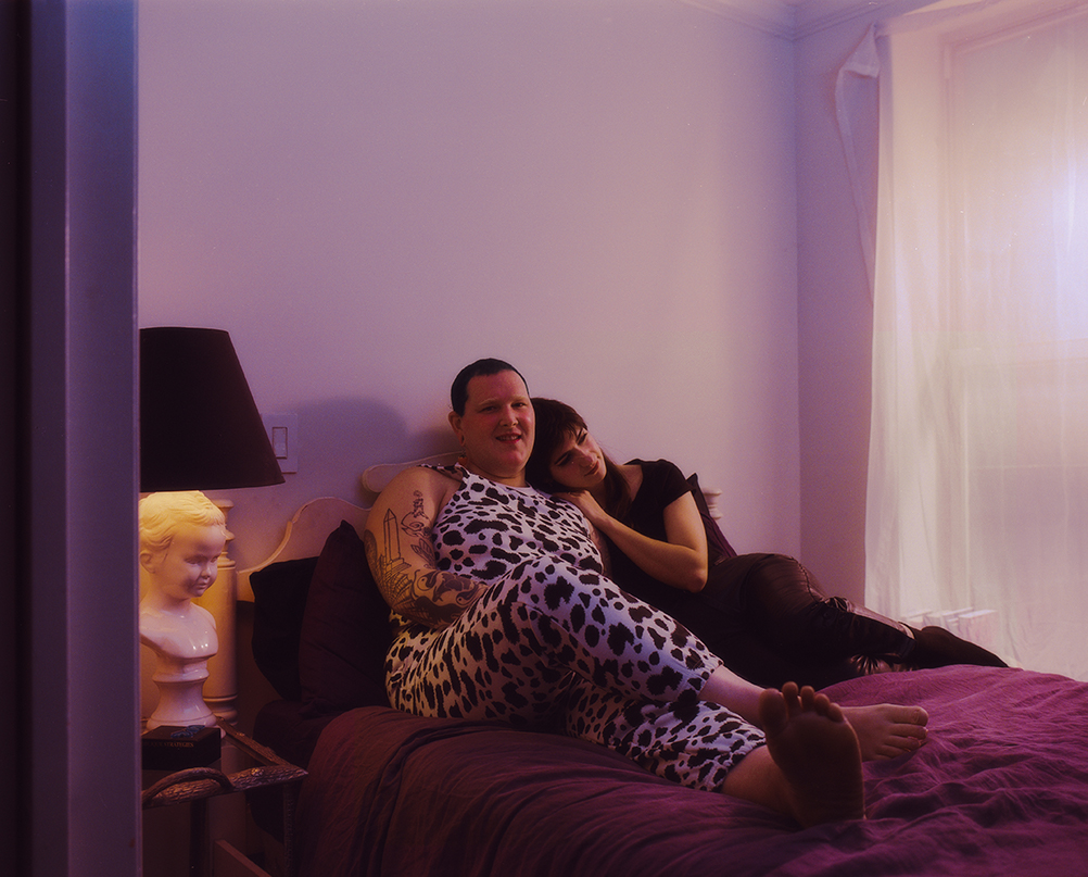 Tell and Camille cuddling in their bed with purple sheets. Telly wears a cow print top and pants and Camille wears all black. 