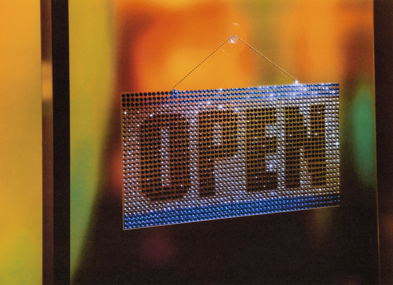 A blue and white sign with OPEN written in all black capitalized letters hangs in the window of a glass door, the sign is made of rhinestones.