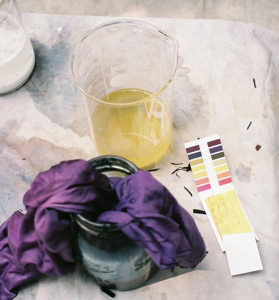 A strip of paper with color swatches sits next to a small jar containing a purple-dyed textile as well. In the frame is also a glass beaker with yellow liquid inside. 