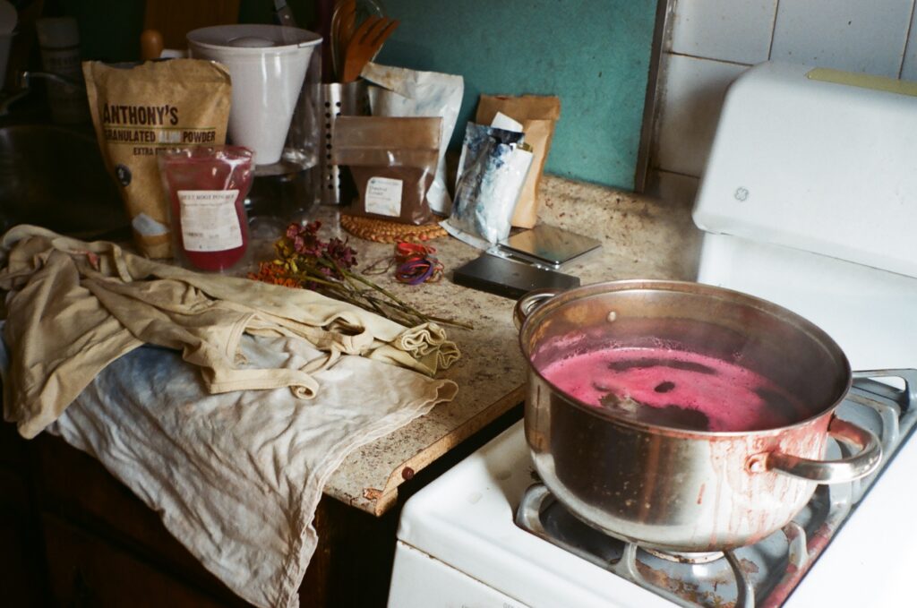 A metal pot with steaming pink liquid sits on a stovetop. On the countertop are a series of powdered alums and dye ingredients as well as a wet white tank top.