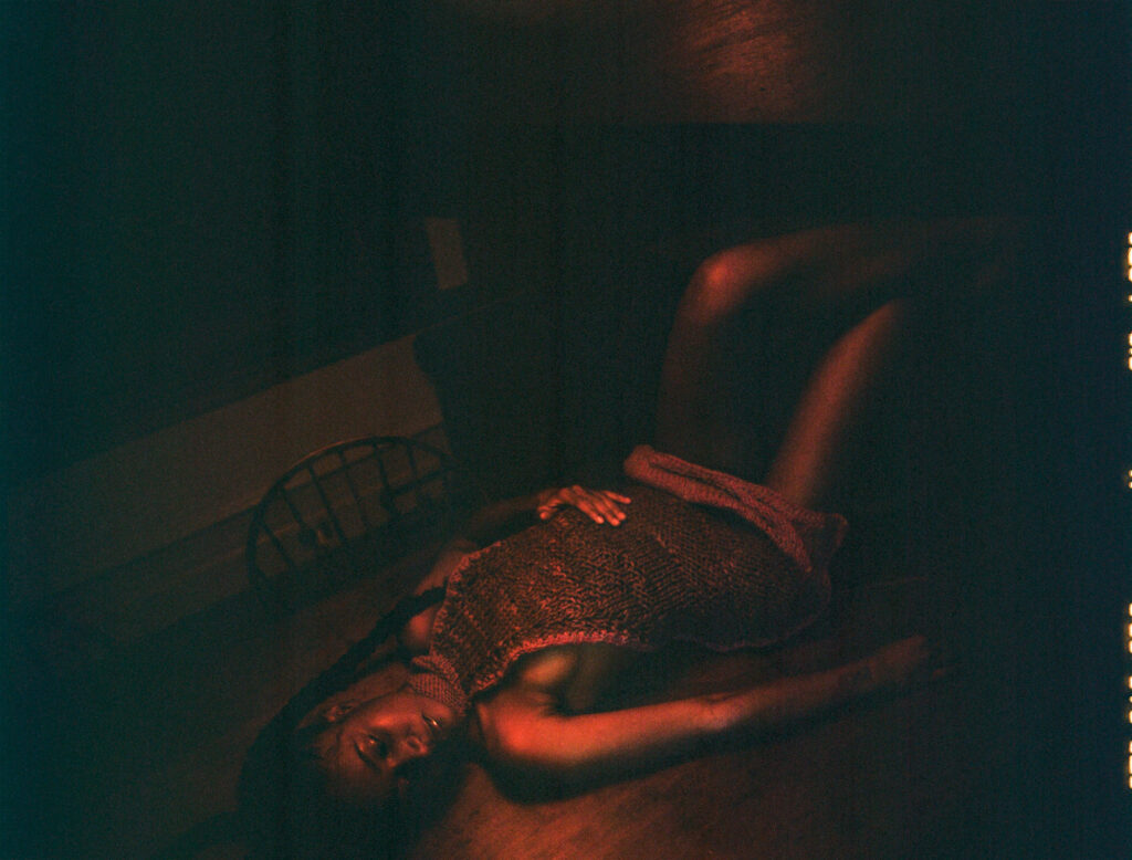 A woman wearing a knit halter-neck apron lies on the ground with her legs propped up on a chair. The photo is taken in dark, red-orange lighting