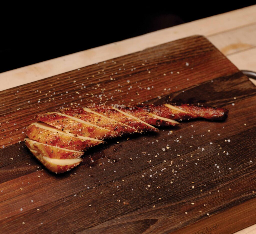 Photo of a cooked fillet of meat sliced expertly on a wooden cutting board with seasoning sprinkled on. 