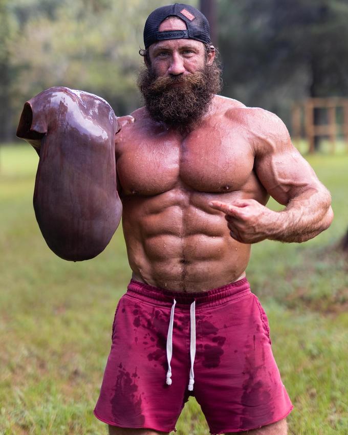 A shirtless man with bulging muscles holds a large cow's liver in one hand pointing his hand toward it with the other. He wears a backwards baseball cap and swim trunks.