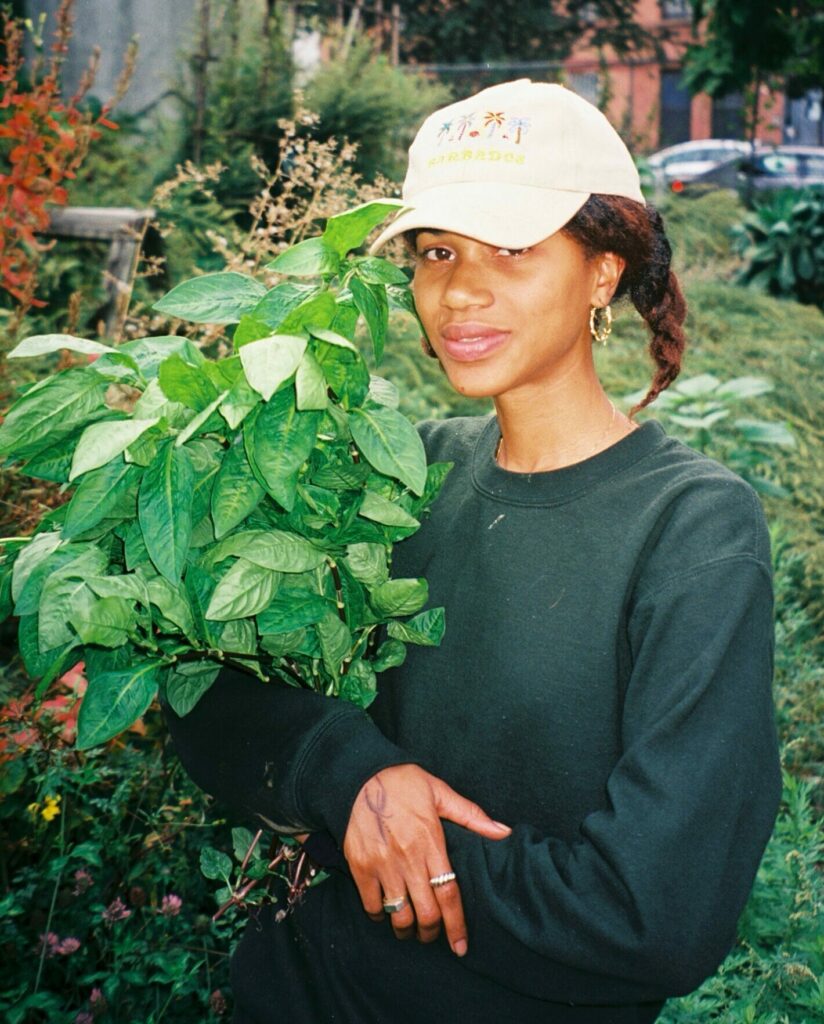 A girl wearing a green sweatshirt and a white baseball cap with the words Barbados embroidered across stands in an urban garden cradling a bushel of herbs.