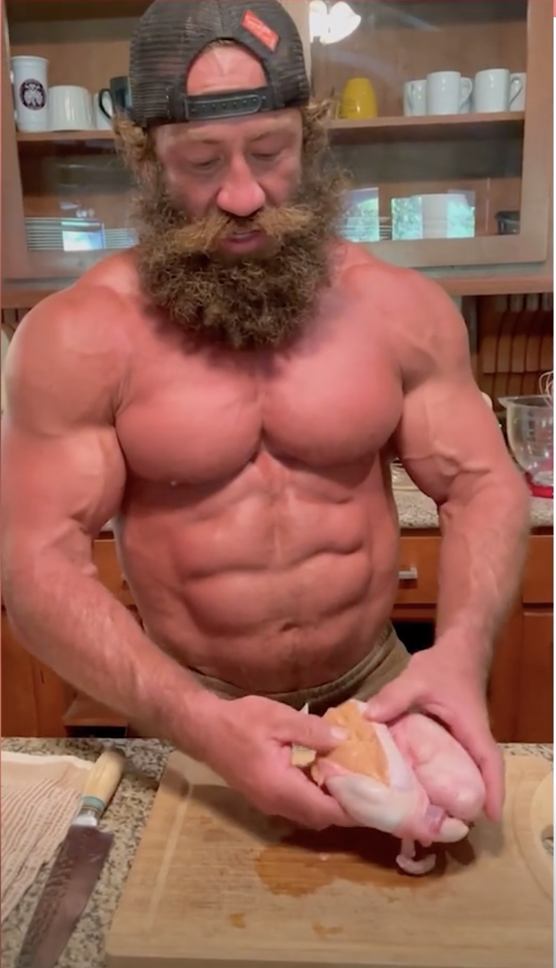 A shirtless man in a kitchen holds raw bull testicles in his hands above a wooden cutting board.