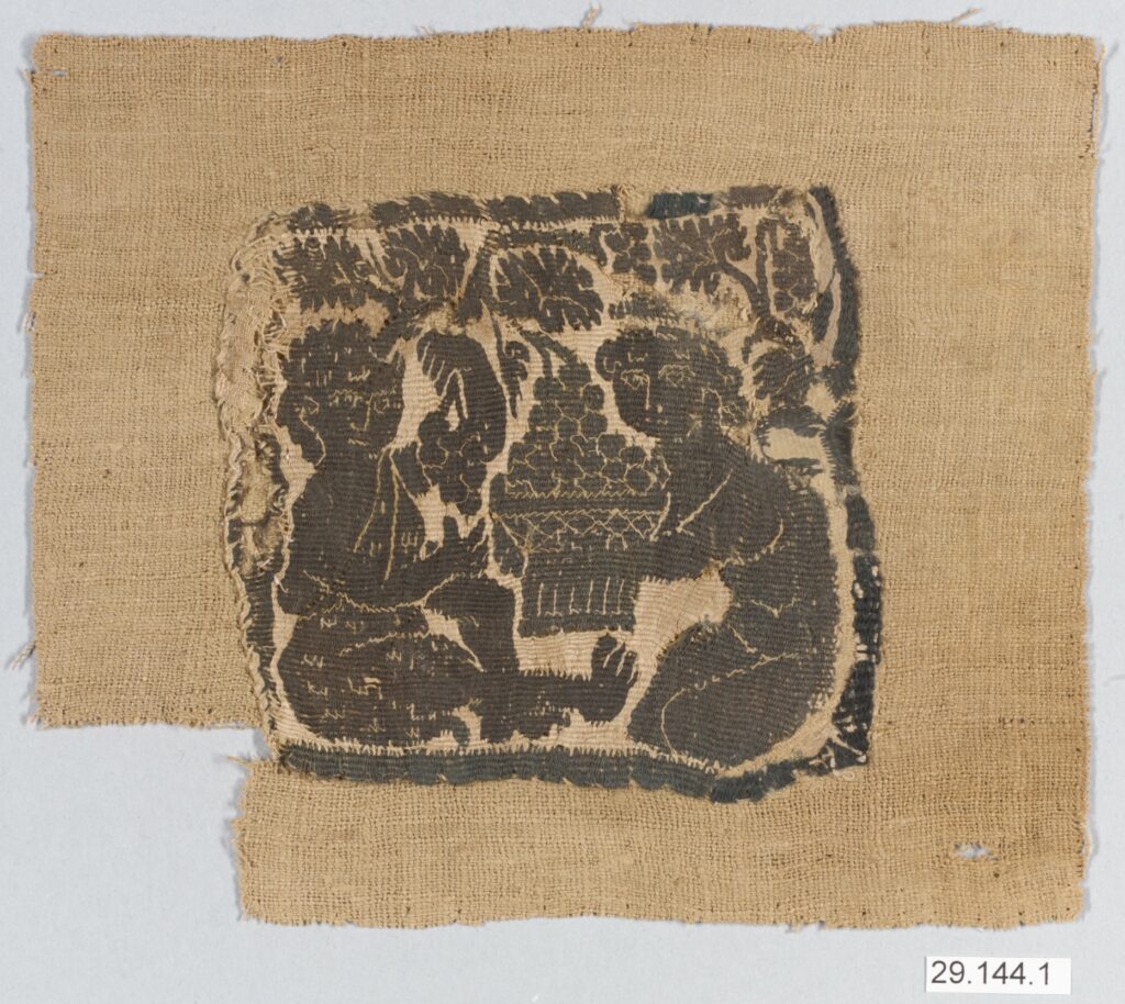 On a piece of linen fabric a black block-cut print of two individuals sitting below a grape vine harvesting grapes. 