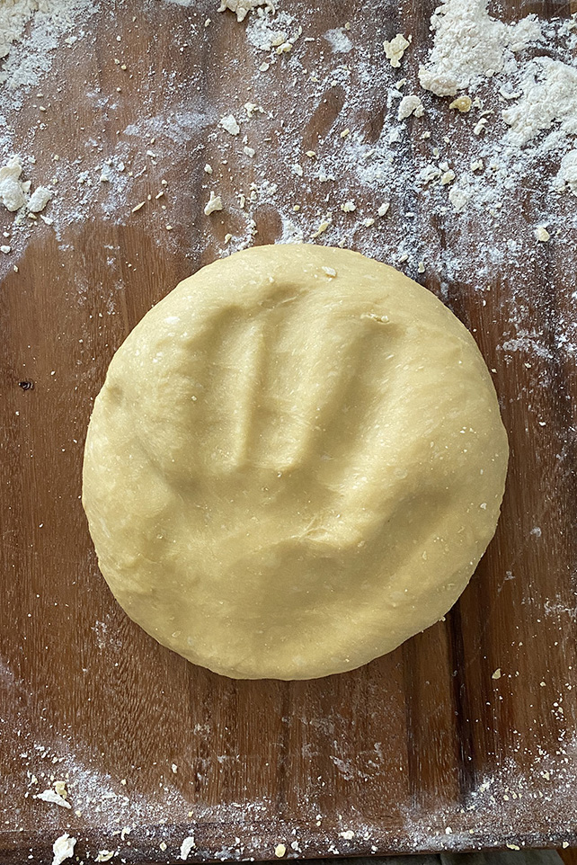 A ball of pasta dough with a hand print on top. The ball of pasta dough is on a flour-dusted wooden table.