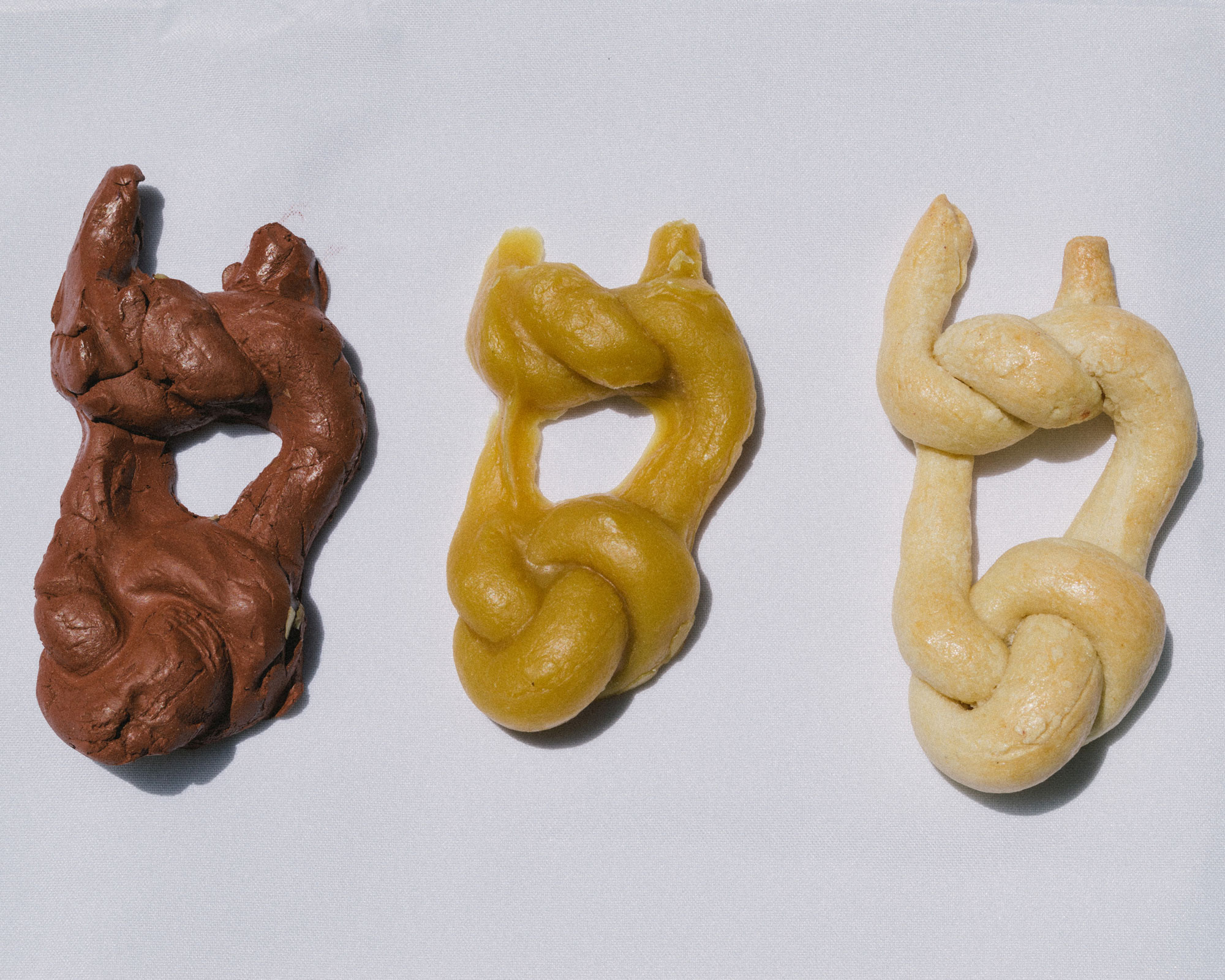 Three bread sculptures are arranged in a horizontal line. Each sculpture is identical and in the shape of two knots. They are arranged in ascending color order from dark brown, to yellow beige, to white beige.