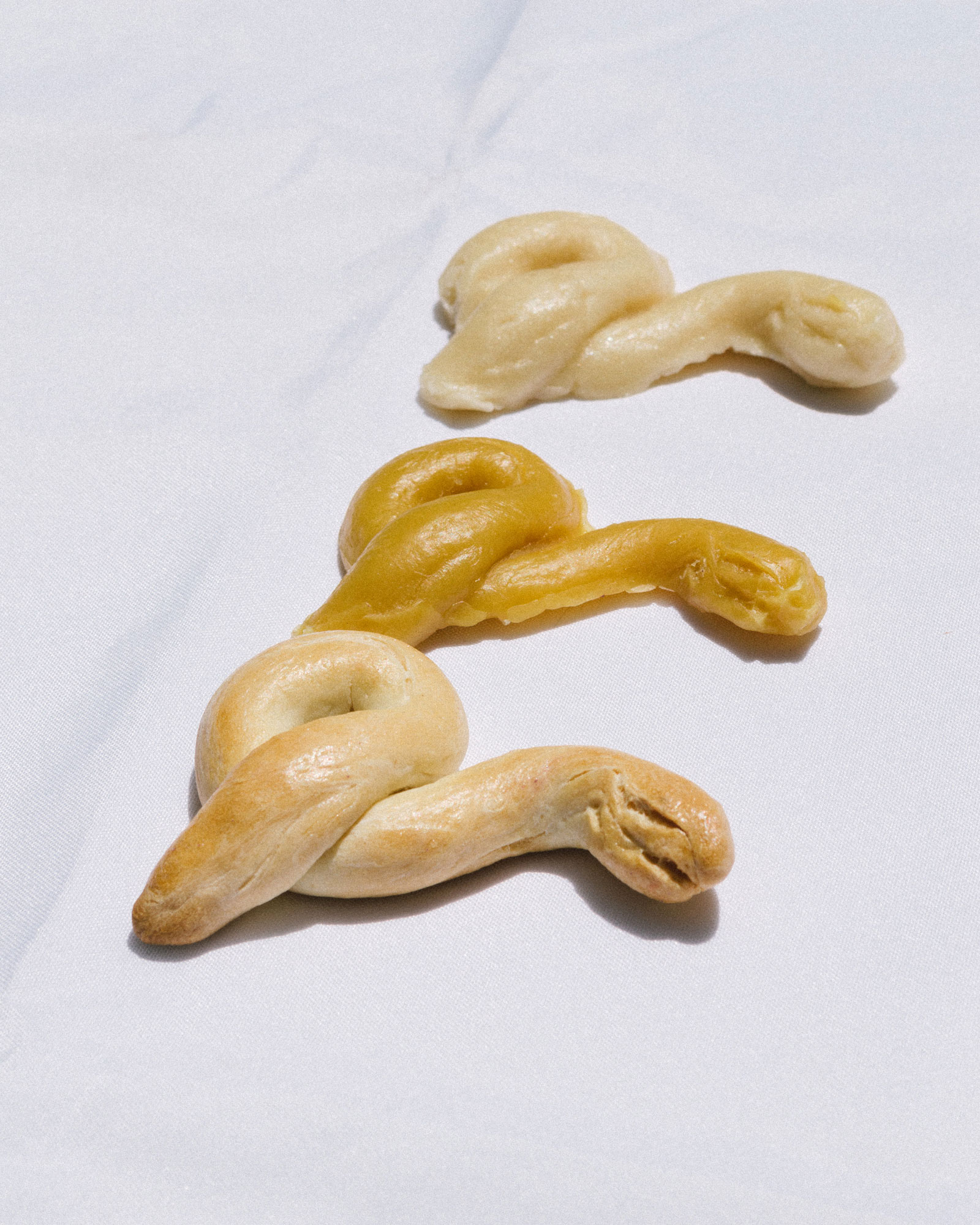 Three bread sculptures are positioned in a vertical line. Each sculpture is identical— a snake head who's body curls over itself once to form a loop in the back. The sculptures are bread-colored, yellow and a pale beige, arranged in order of baked-ness.