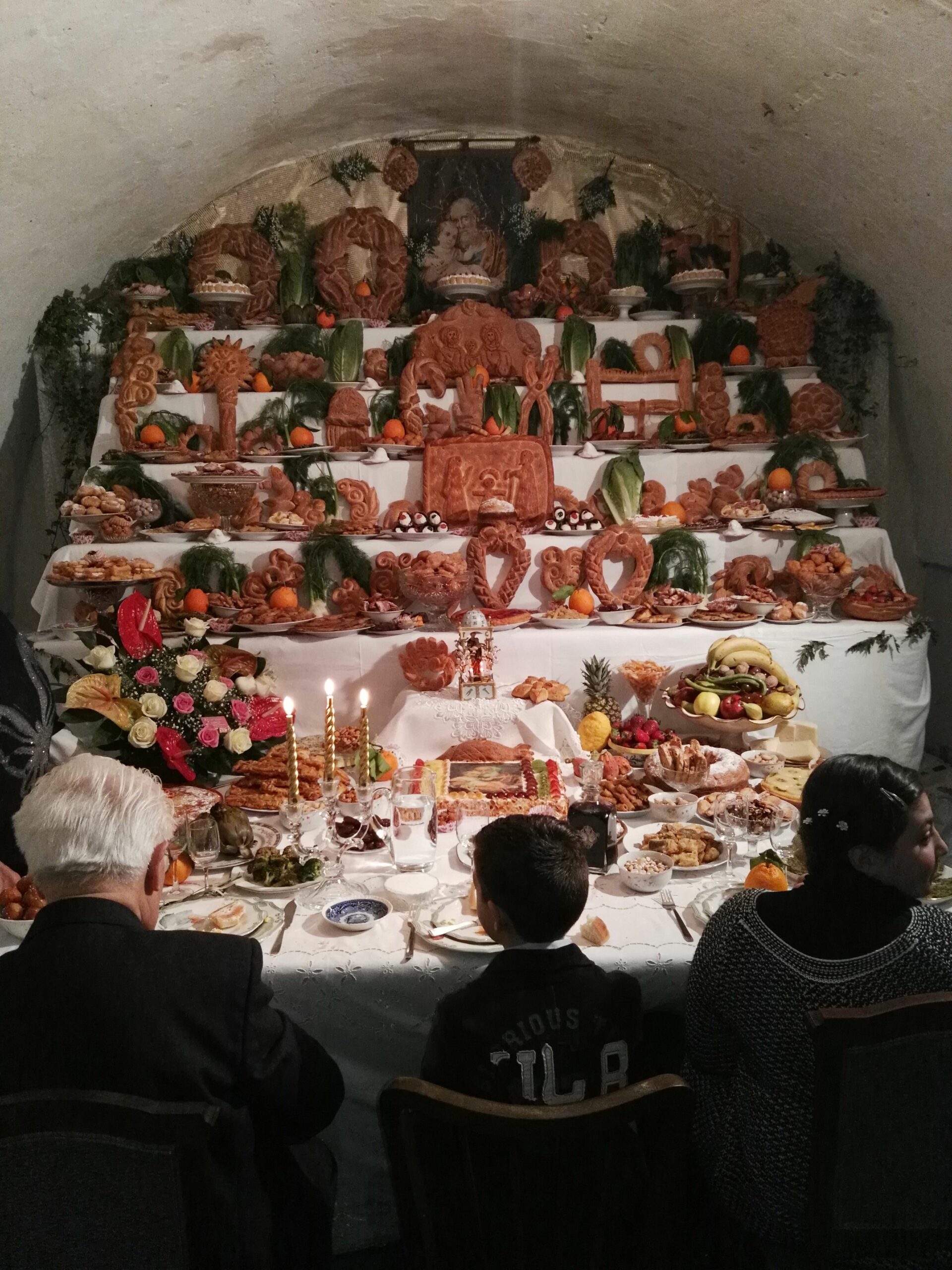 Inside a white curved room is an altar with five layers of shelving, each layer is stacked with objects fashioned out of bread (animals, housing, etc.) Interspersed between these bread objects are fruits and vegetables. Candles are lit, three individuals sit at a dining table facing the alter, an elderly man on the left, a small child in the middle, and another, older child whose head is turned to someone off camera.