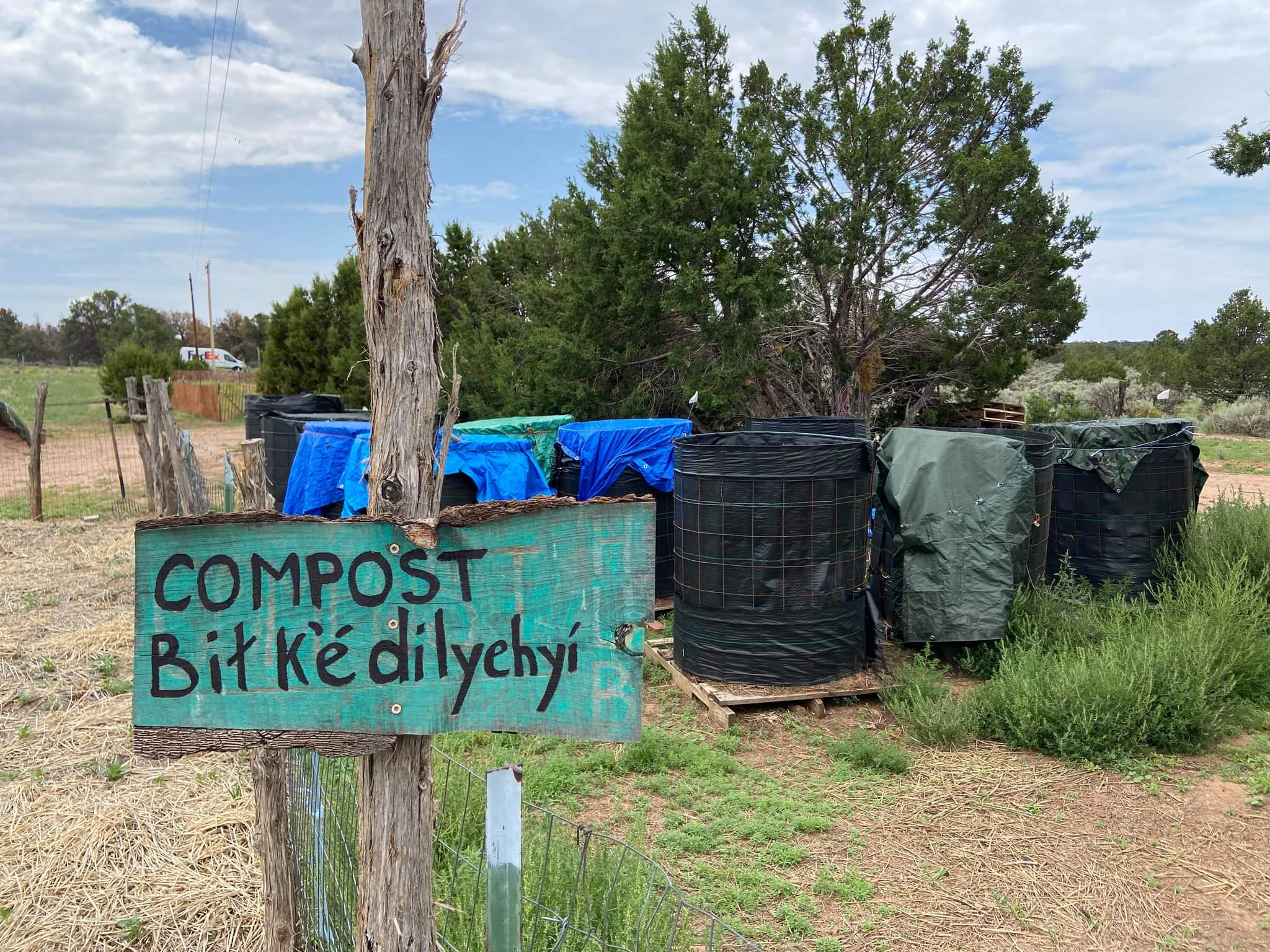 Several compost bins wrapped in cobalt blue, black, and olive green tarps sit below a blue sky with clouds, in front of a smattering of trees. A fence with a green sign reading Compost and Bit k'e'dilychyi is in the foreground.