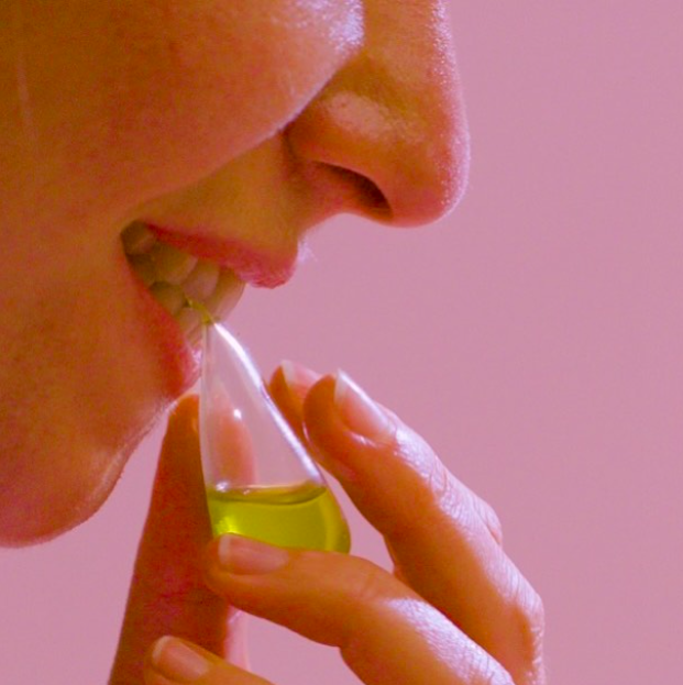 A close up of a person's face against a pink background shows them holding a pipette with green liquid up to their mouth