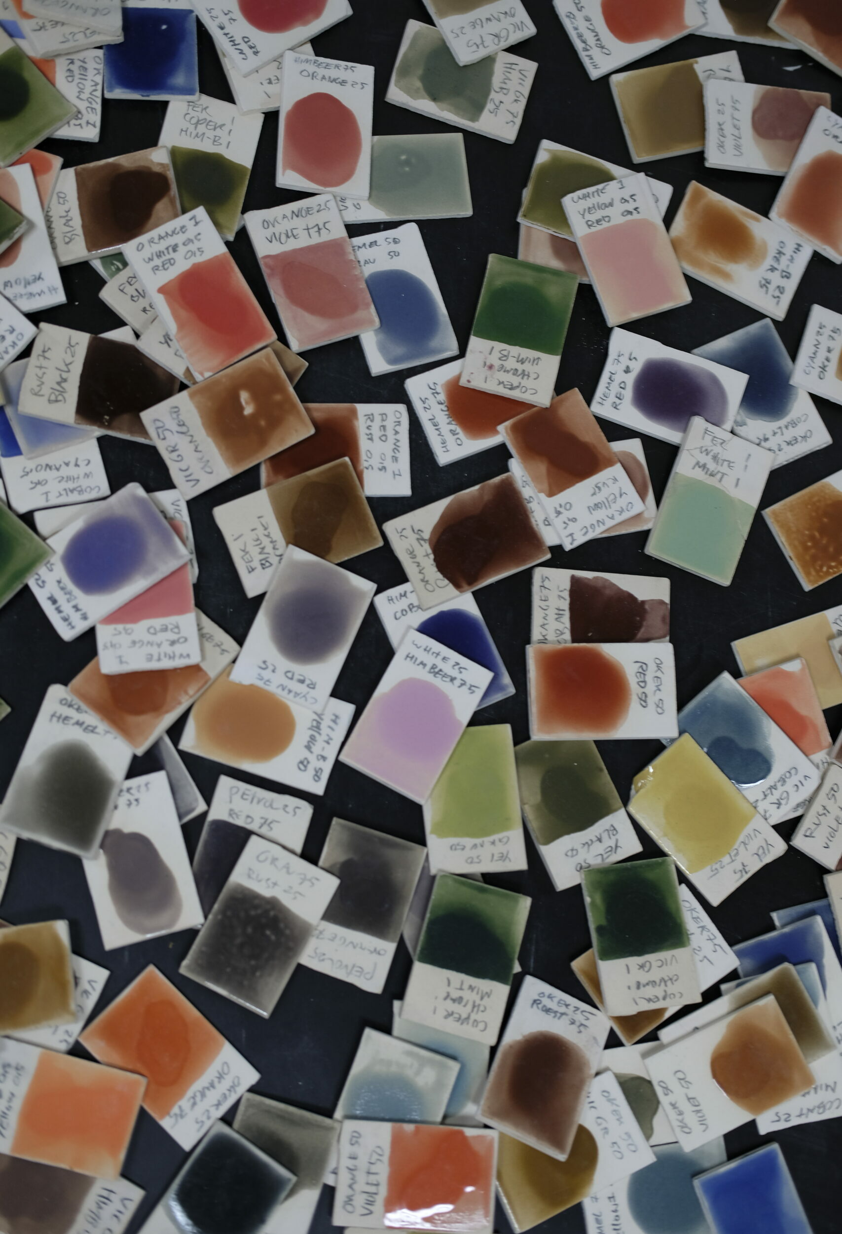 Dozens of small cards with glaze samples in various colors are scattered on a black table. Each card has a description of the glaze number and name.