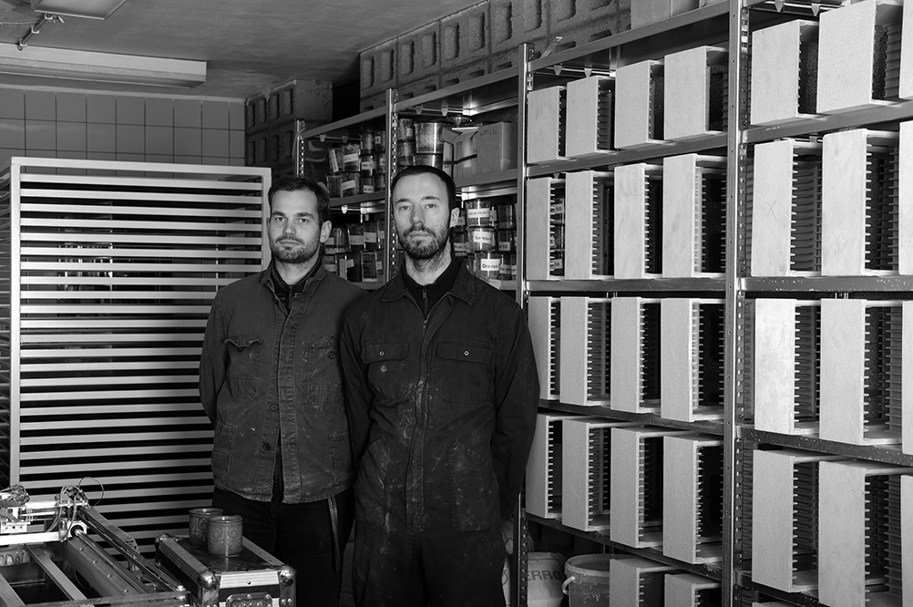 In a black and white photo two men of the same height stand together looking into the camera with serious expressions and with their hands behind their backs. Behind him are racks where ceramic tiles are stacked.