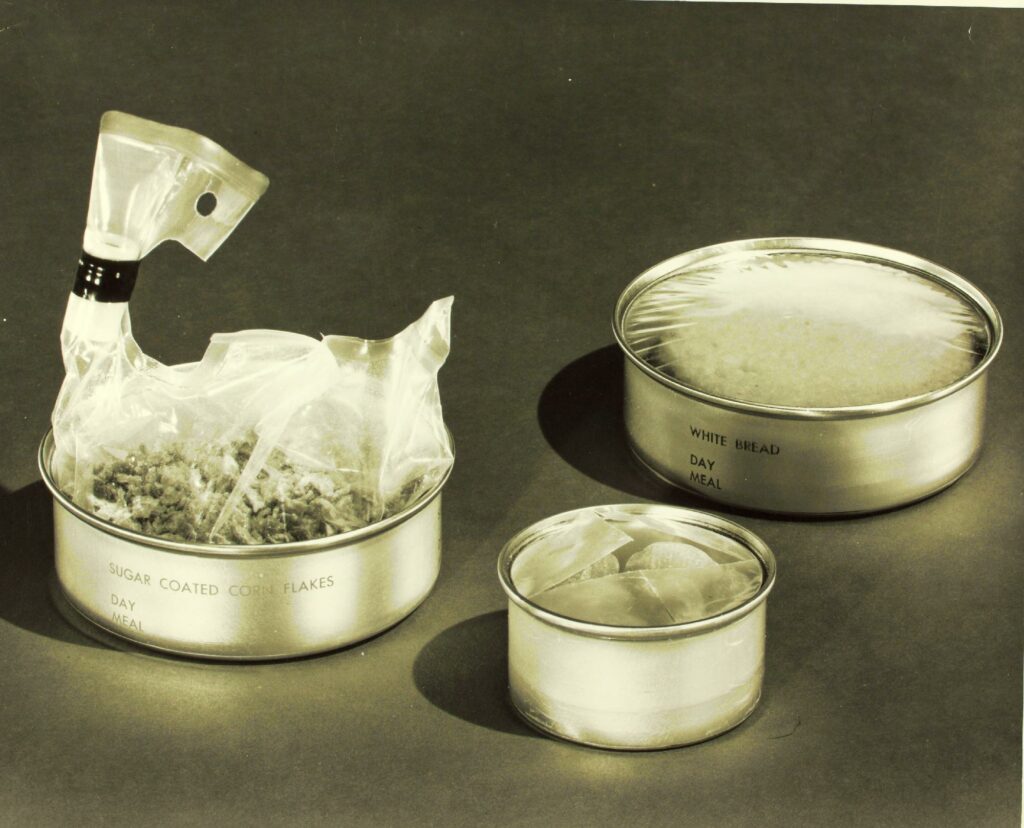Three aluminum cans of space food, each labeled with what is contained within it (i.e. white bread)