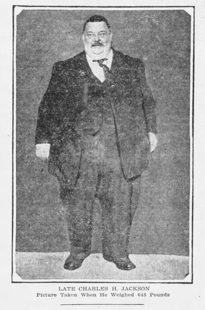 A large man in a suit stands in a black and white photograph. At the bottom the following is inscribed "Late Charles H. Jackson. Picture taken when he weighed 648 pounds."