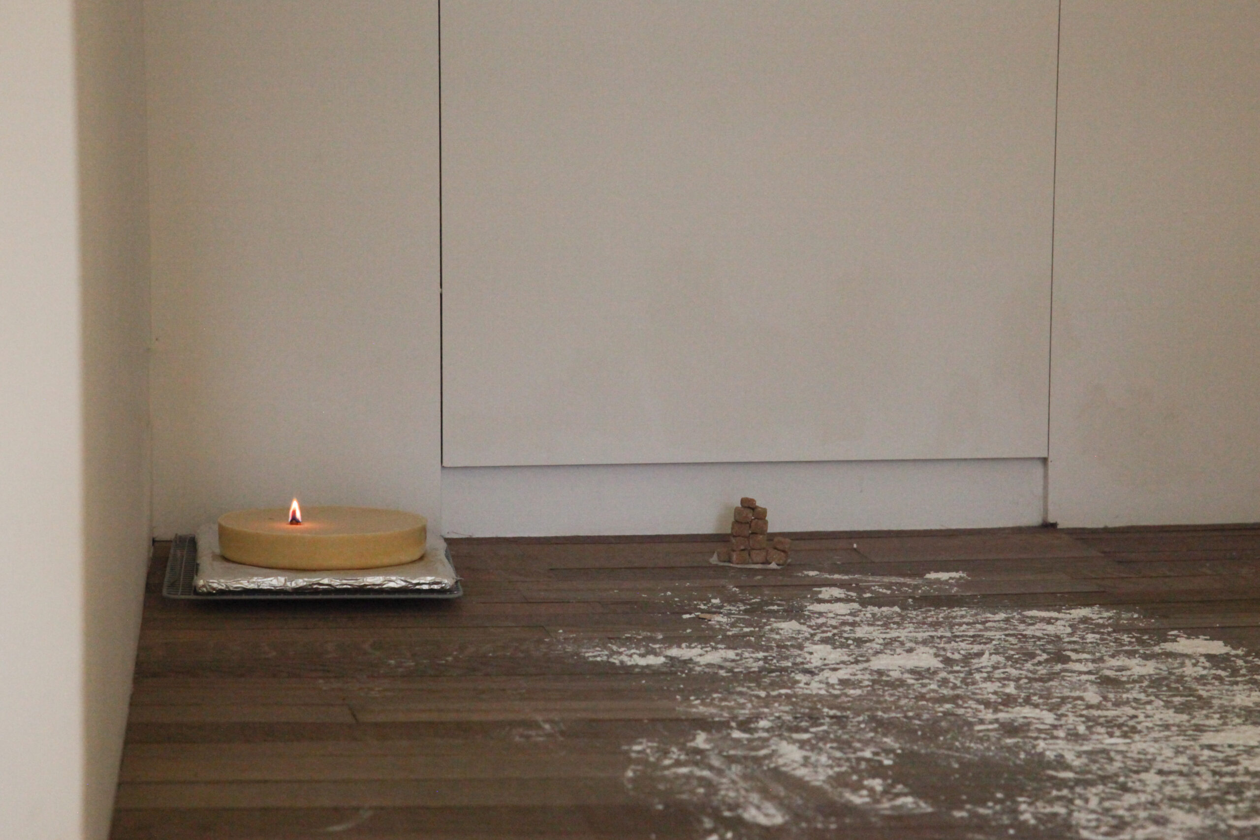 A room with white walls and brown hardwood floors. In the corner is a cake-shaped candle on top of an aluminum foil-wrapped tray. A small flame is lit. Off to the right is a small stack of brown sugar cubes which form a tower. Flour is dusted across the floor.