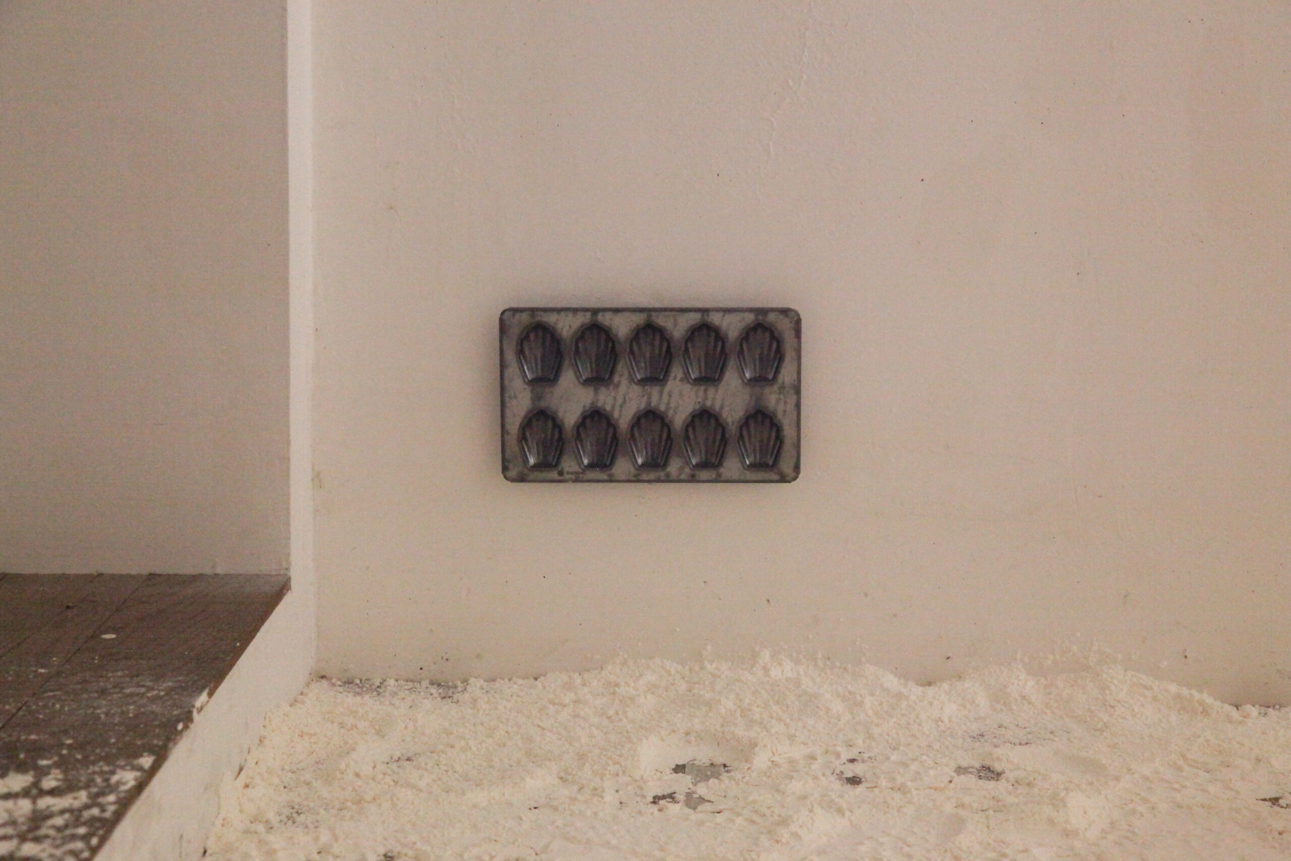 A silver tray mold for ten madeleine cookies is hung up on the wall slightly above the ground. Below on the ground the floor is covered in a layer of flour.