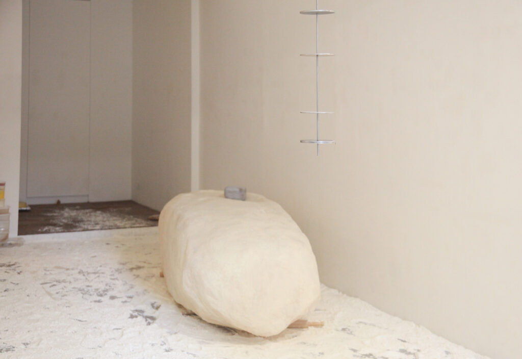 A white, rounded rock-shaped sculpture made of flour sits in a white room with flour scattered on the floor. A smaller grey rock sits on top of the sculpture