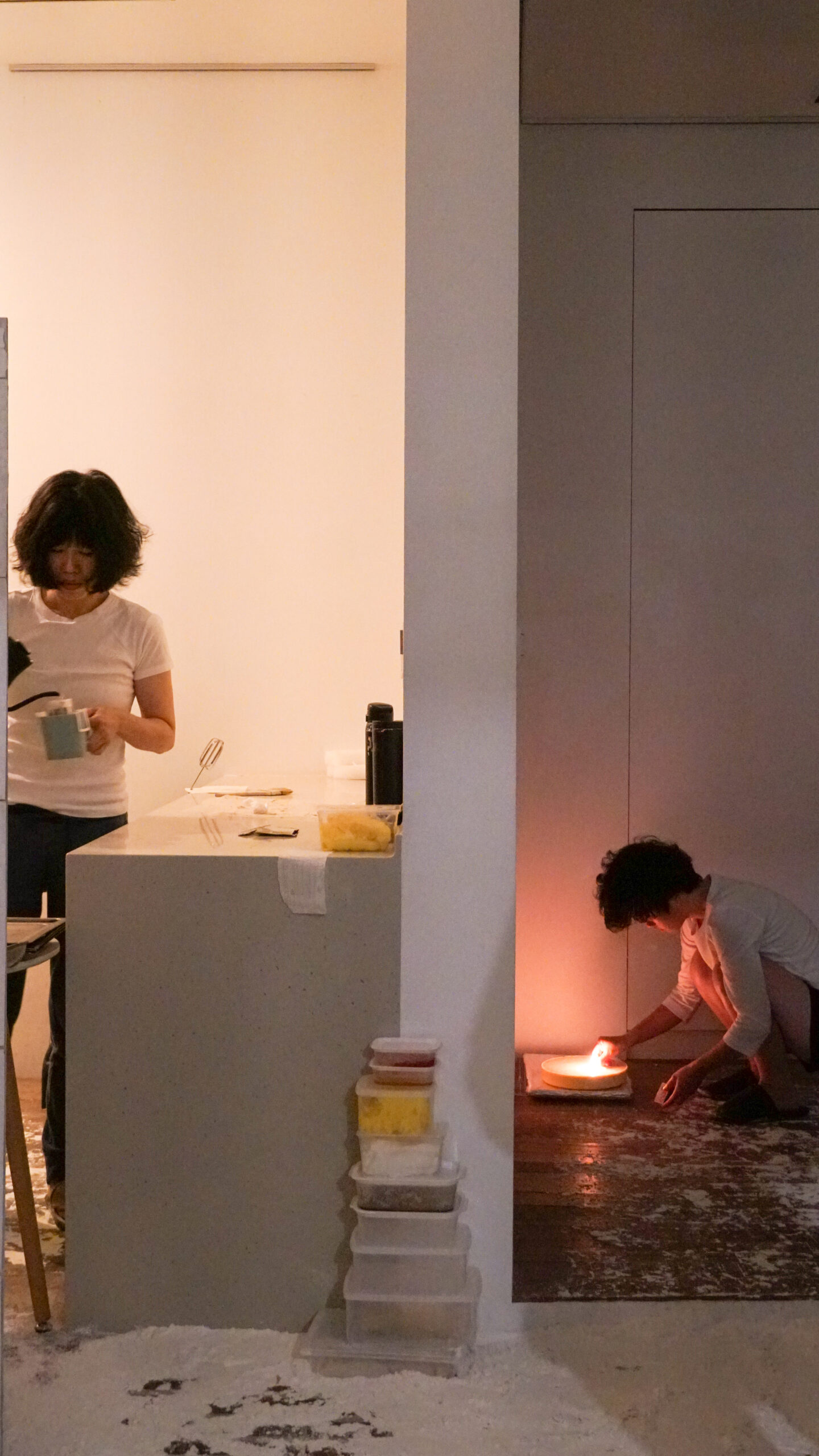Two individuals are shown installing the exhibition. On the left petite femme stands to the left pouring water from a kettle into a cup. Separated by a wall, another figure is seen crouched installing another work of art in a dimmer room, the light from the artwork casts a warm glow.