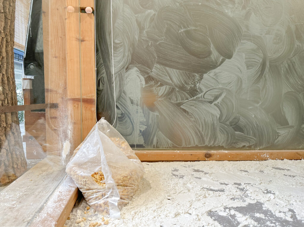 A sack of wheat is shown propped against the inside window of a bakery. A layer of flour has been sprinkled across the floor. One of the adjacent windows is dirty with residue from circular wiping motions.