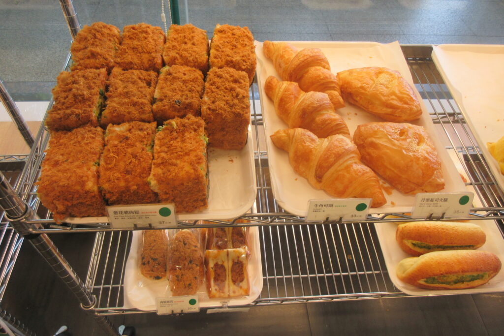 Four trays of baked goods sit on the rack in a bakery. Croissants, buns dusted in pork floss and rolls. 