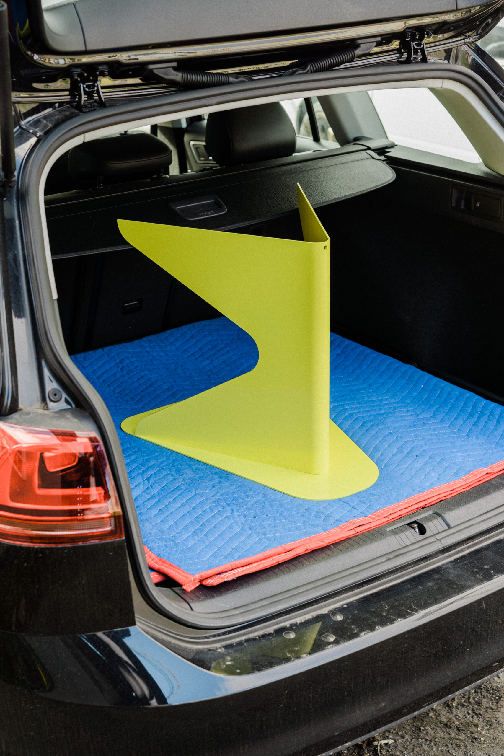 A lime green bent metal stoll sits upright in the trunk of a car atop of a blue moving blanket with orange trim.