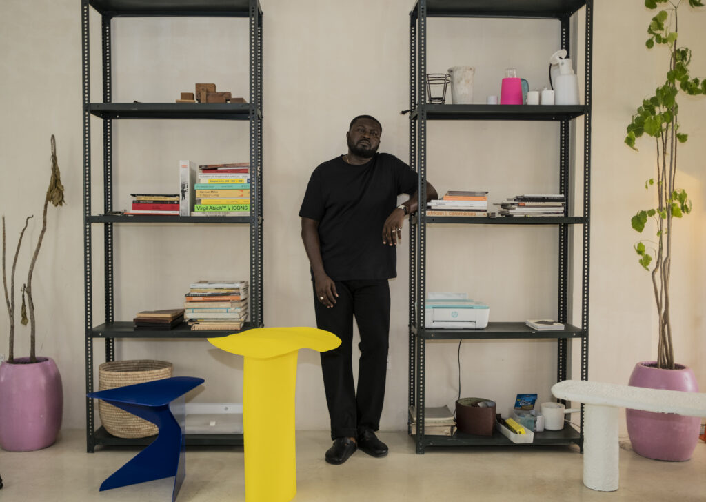 Nifemi dressed in all black stands in between two metal bookcases. He leans on one with his elbow. Surrounding him are stools and furniture he's made. A yellow side table, a cerulean blue stool, a white bench, flamingo pink plantholders.
