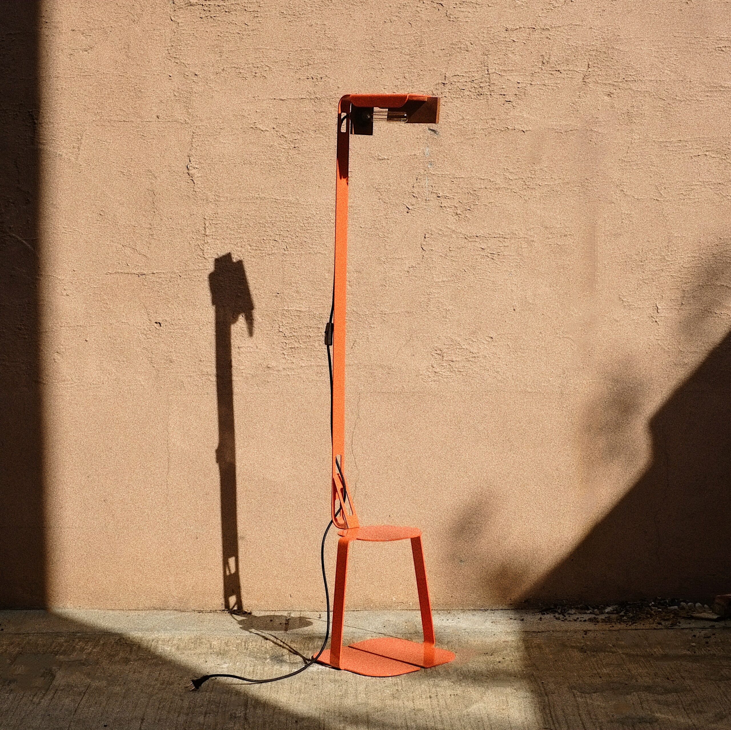An orange metal stool sits on concrete outdoors. A pole extends from the stool, and ends in an overhang which contains a lightbulb.