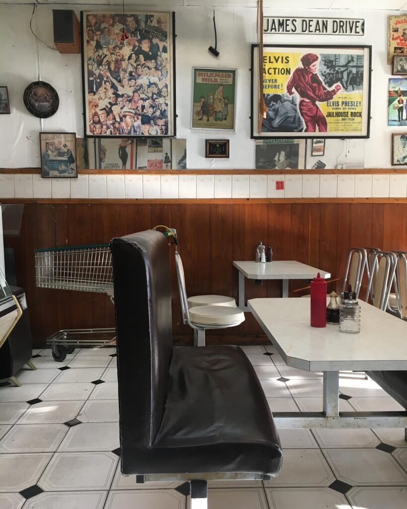 An image of the inside of a greasy spoon diner. A black, free-standing booth sits in the foreground a formica table at it's center. The space has rich wood paneling, and the walls are covered with photos, posters, and memorabilia.