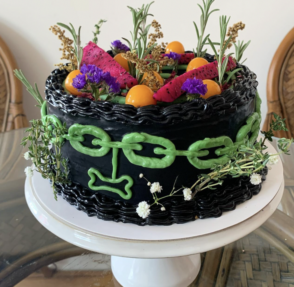 A black cake on a white stand with a green frosted chain decorating the side. Topped with kumquats and amaranth flowers.