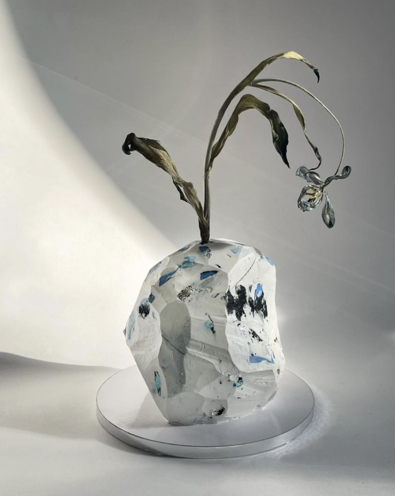 A cake shaped like a rock-like iceberg. White, angular, with splotches of black and blue. A dried flower bends out from the top of the cake, much like a vase. 