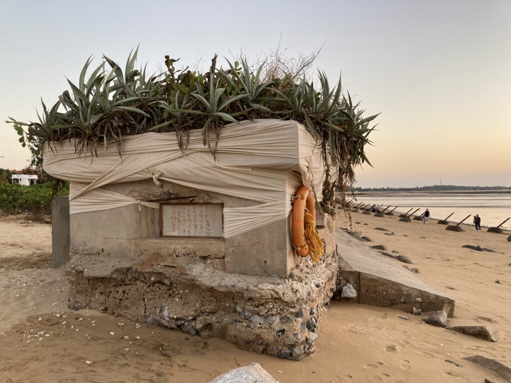 A concrete shrine on a sandy beach is wrapped in diaphonous cream colored cloth. Atop there are mounds of native plans, spiky, they overflow beyond the roof. 