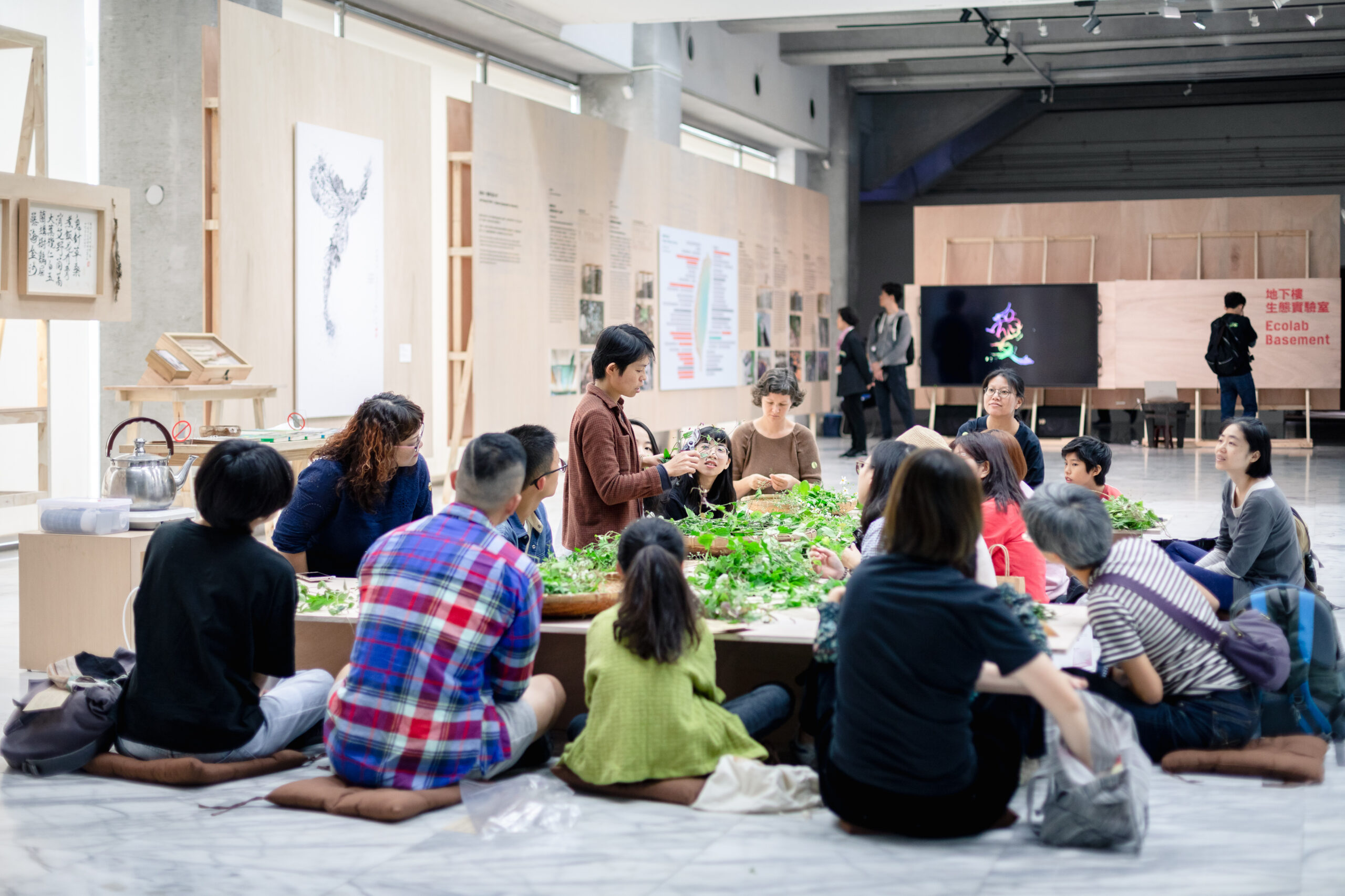 In a light wood paneled art space an individual in a brown shirt holds a handful of leaves in front of a dozen people sitting on floor cushions around a table covered with mounds of foraged greens.