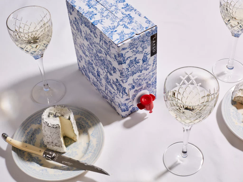 Three crystal goblets filled with white wine sit around a box of wine that is blue china patterned. A small plate with a circular soft cheese sits next to a sharp cheese knife. The table is covered in white tablecloth.