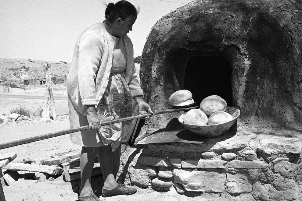 An woman scoops baked bread out of a horno, an oven made of earth that rises from a rocky bed. 
