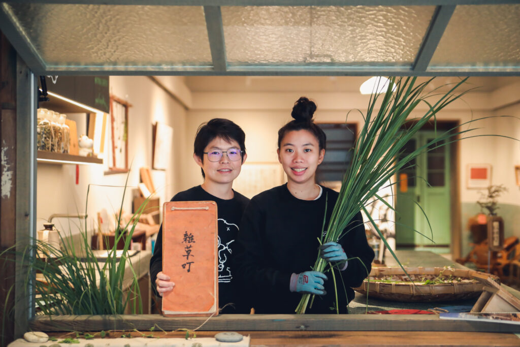 Two individuals dressed in black sweaters stand inside of a glass window one holding grasses and the other a booklet. 